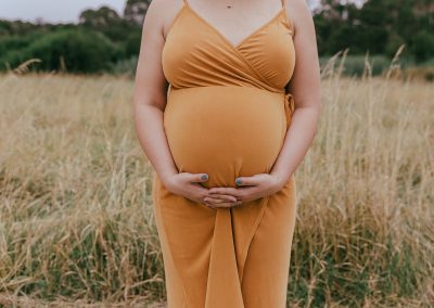 Pregnant lady standing in a grass field. She's wearing a mustard dress and holding her hands below her belly. Only her chest down to her knees can be seen